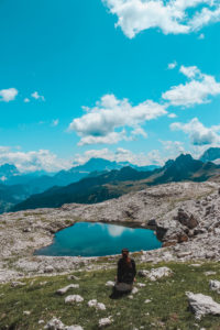 piccola lago alpino verde smeraldo circondato dalle alte vette dei monti. Una ragazza seduta di spalle guarda il panorama