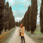 ragazza con cappotto e cappello beige tra filari di cipressi in Toscana
